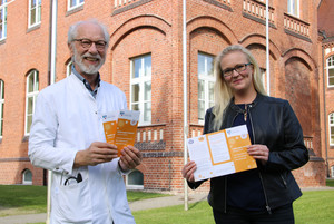 Zwei Personen stehen vor einem Backsteingebäude und halten Fyler in den Händen.