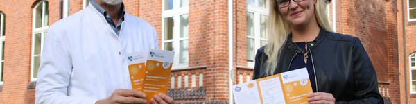 Zwei Personen stehen vor einem Backsteingebäude und halten Fyler in den Händen.