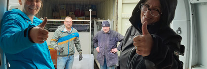 Mitglieder des Vereins Hand in Hand stehen am Container mit den eingeladenen Tafeln und schauen in die Kamera.