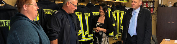 Vier Personen stehen in einem Raum und unterhalten sich. Im Hintergrund hängen Feuerwehruniformen.