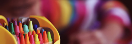 Eine Box mit bunten Stiften, im Hintergrund malt ein Kind.