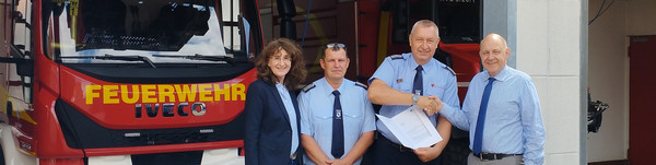 Vier Personen stehen vor einer Feuerwehr-Halle, in der zwei Feuerwehren stehen - Klick öffnet Bildbetrachter