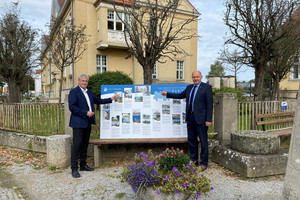 Zwei Männer stehen vor einem Gebäude, zwischen ihnen steht auf einer Bank die Schautafel.