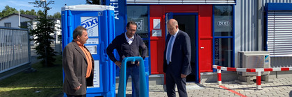 Drei Männer stehen vor einer Dixi-Toilette und unterhalten sich.