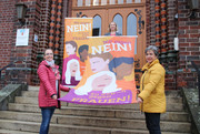 Drei Frauen stehen auf einer Treppe und halten eine orange Fahne mit der Aufschrift "Wir sagen Nein! zu Gewalt gegen Frauen".