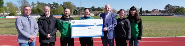 7 Personen stehen auf der Laufbahn im Premnitzer Stadion und die 3 in der Mitte halten den symbolischen Spendenscheck. - Klick öffnet Bildbetrachter