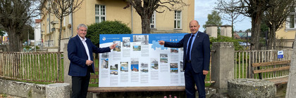 Zwei Männer stehen vor einem Gebäude, zwischen ihnen steht auf einer Bank die Schautafel.