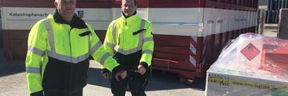 Zwei Männer stehen vor einem Container. Sie bedienen einen Hubwagen mit Materialien.