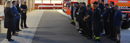 Vier Personen stehen vor einer Gruppe von Feuerwehrleuten, im Hintergrund stehen verschiedene Feuerwehrfahrzeuge.