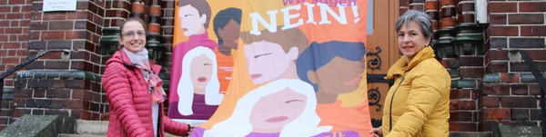 Drei Frauen stehen auf einer Treppe und halten eine orange Fahne mit der Aufschrift "Wir sagen Nein! zu Gewalt gegen Frauen".