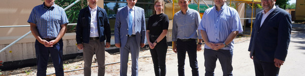 Eine Gruppe von sieben Menschen steht vor einem Baugerüst, das vor der Schule steht. An der Fassade ist ein Wandbild zu erkennen.