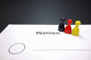 Auf einem Blatt mit der Aufschrift "Wahlen" stehen drei Spielfiguren in den Farben Schwarz, Rot und Gelb..