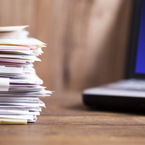 Large stack of files, paperwork.  Desk, office, computer.