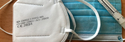 Auf einem Tisch liegen zwei Mund-Nasen-Bedeckungen sowie ein Corona-Test.