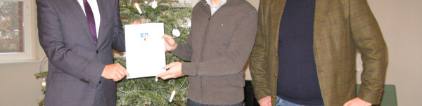 Drei Männer stehen an einem Weihnachtsbaum, zwei halten eine Dokumentenmappe zwischen sich.