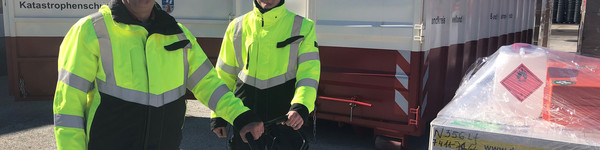 Zwei Männer stehen vor einem Container. Sie bedienen einen Hubwagen mit Materialien.