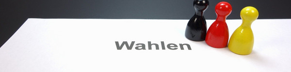 Auf einem Blatt mit der Aufschrift "Wahlen" stehen drei Spielfiguren in den Farben Schwarz, Rot und Gelb..