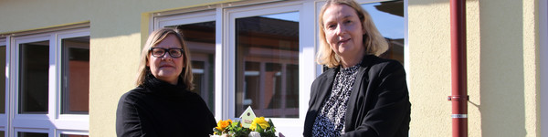 Zwei Frauen stehen vor einem Gebäude und halten zwischen sich eine Schale mit Blumen.