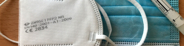 Auf einem Tisch liegen zwei Mund-Nasen-Bedeckungen sowie ein Corona-Test.