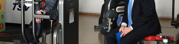 Landrat Lewandowski und Birgit Faber sitzen auf Sportgeräten in einem Fitness-Raum und unterhalten sich..