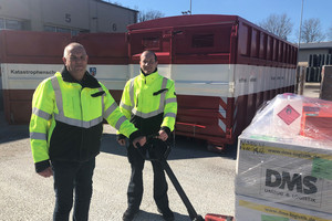 Zwei Männer stehen vor einem Container. Sie bedienen einen Hubwagen mit Materialien.