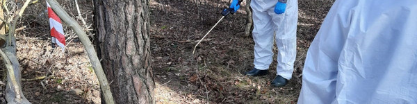 Zwei Personen in weißen Schutzanzügen üben die Desinfektion eines möglichen Fundortes in einem Waldstück.