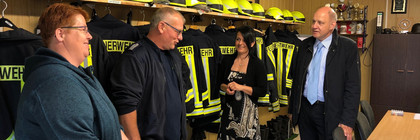 Vier Personen stehen in einem Raum und unterhalten sich. Im Hintergrund hängen Feuerwehruniformen.