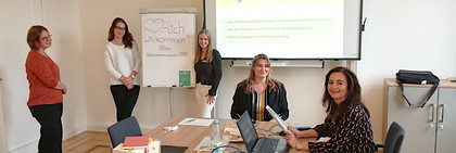 Fünf Frauen sitzen und stehen verteilt in einem Raum.