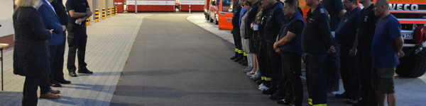 Vier Personen stehen vor einer Gruppe von Feuerwehrleuten, im Hintergrund stehen verschiedene Feuerwehrfahrzeuge.