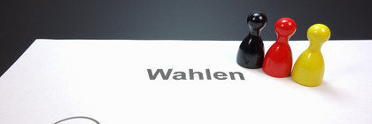Auf einem Blatt mit der Aufschrift "Wahlen" stehen drei Spielfiguren in den Farben Schwarz, Rot und Gelb..