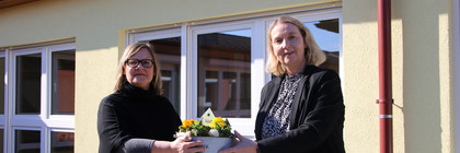 Zwei Frauen stehen vor einem Gebäude und halten zwischen sich eine Schale mit Blumen.
