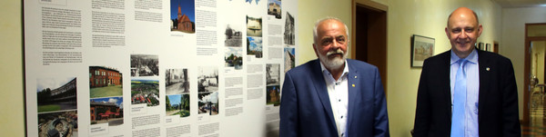 Zwei Männer stehen in einem Flur, an der Wand hängt eine Schautafel.