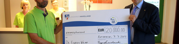 Dr. Wilke und Landrat Lewandowski stehen in der Arztpraxis und halten einen symbolischen Scheck zwischen sich.